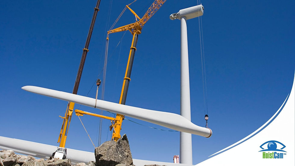 A large white wind turbine blade is being lifted by two yellow cranes against a clear blue sky. The blade is long and slender, with one end elevated and the other resting on the ground or a support structure. Rocks are visible in the foreground, and the base of a wind turbine tower is also present. In the bottom right corner, there's a blue and white "HoistCam" logo.
