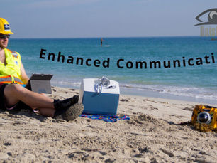 A man wearing a yellow hard hat, sunglasses, and a reflective safety vest sits on a small blue beach chair on a sandy beach. He is holding a drink in his left hand and has a tablet on his lap. Beside him is a light blue cooler. Further to the right, a yellow and black HoistCam camera is partially buried in the sand with some seaweed. The ocean stretches out behind him under a clear sky, with a person on a paddleboard in the distance. Overlaid on the image in the center is the text "Enhanced Communication" in dark gray. In the top right corner, the "HoistCam" logo is visible.