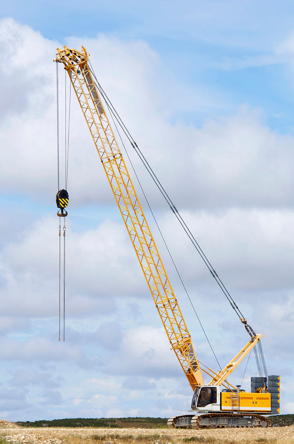 Yellow crawler crane with a long lattice boom and hook, positioned on a construction site under a partly cloudy sky.