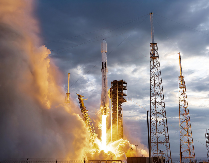 Rocket launching from a space facility with support towers and fiery exhaust against a cloudy sky.