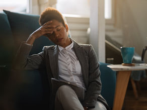 A professionally dressed woman sits on a couch holding a tablet, resting her hand on her forehead with her eyes closed, appearing tired or stressed, with a coffee mug on a nearby table in a softly lit living room.