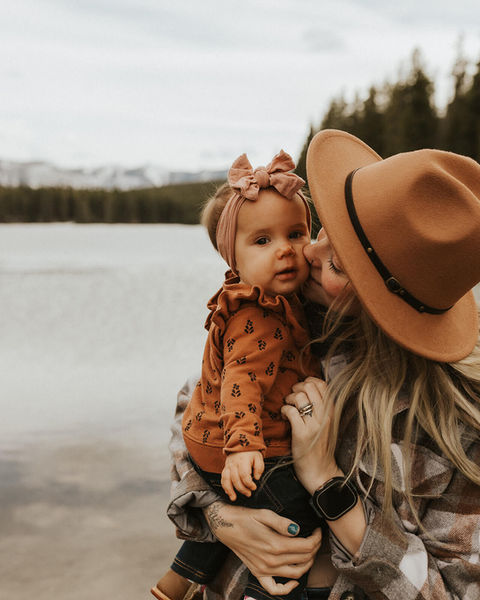 Mother in a knit hat hugging her young daughter in an autumn-colored mountain landscape.