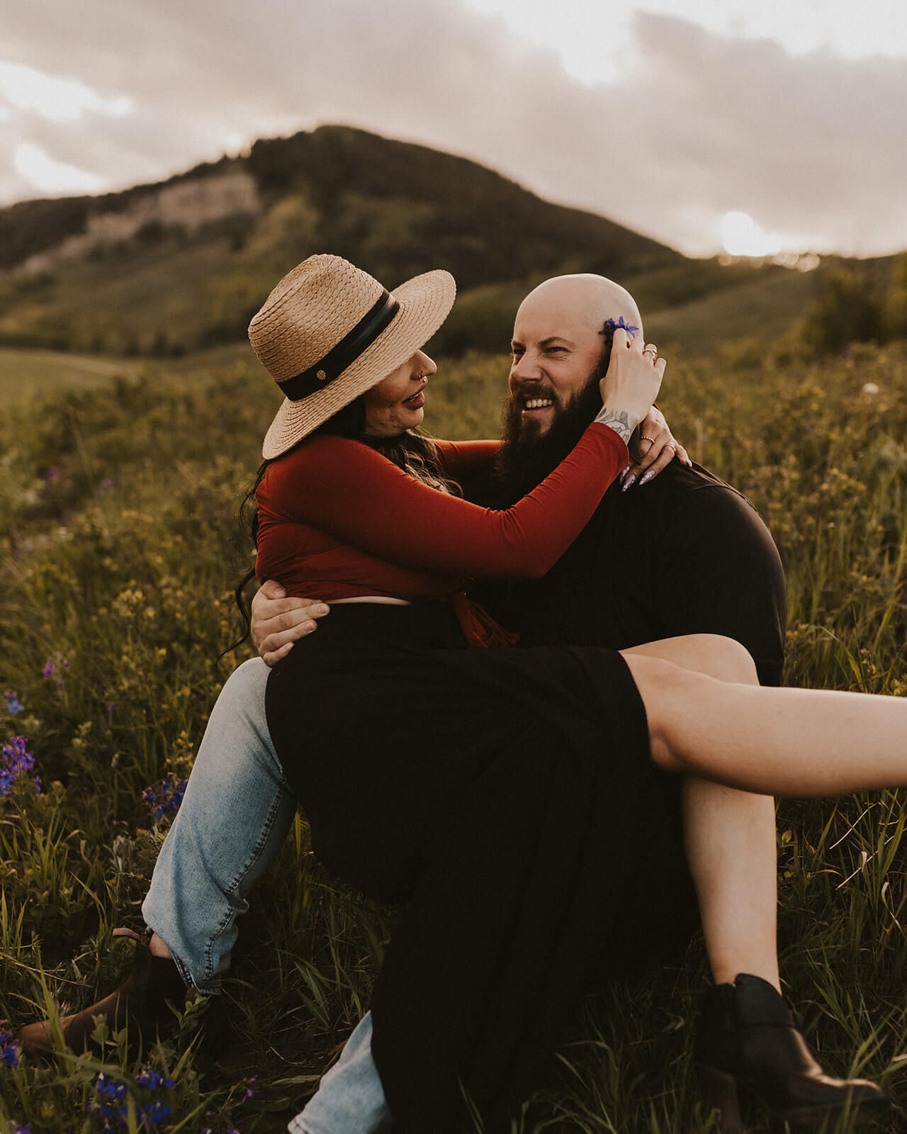 Man tenderly holding woman as they laugh together, dressed in black, with expansive mountain scenery in the background.
