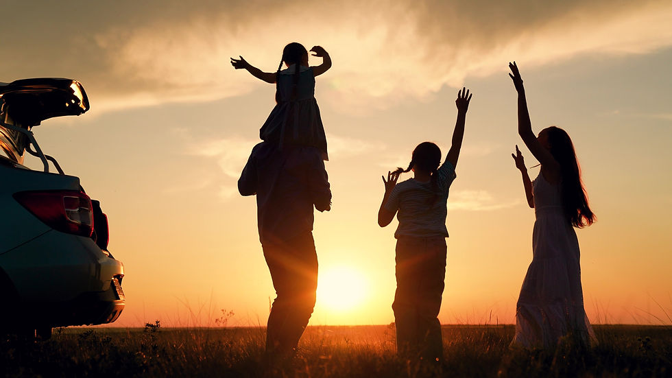 A family silhouetted against a low sun beside an open car boot, arms raised in a field at the end of the day.
