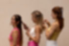 Drei junge Frauen mit langen Haaren stehen hintereinander in einer Reihe und flechten sich gegenseitig die Haare. Sie tragen sommerliche, farbenfrohe Kleidung in Rosa-, Weiß- und Gelbtönen. Jede von ihnen hat eine individuelle Frisur, teilweise mit Haarspangen oder Clips verziert. Die Frau vorne lächelt entspannt, während die anderen beiden konzentriert an den Haaren arbeiten. Der Hintergrund ist eine warme, neutrale Wandfarbe, die das freundliche und harmonische Gesamtbild unterstreicht. Die Szene vermittelt eine fröhliche, unbeschwerte Atmosphäre und feiert Freundschaft sowie gemeinsames Hairstyling.