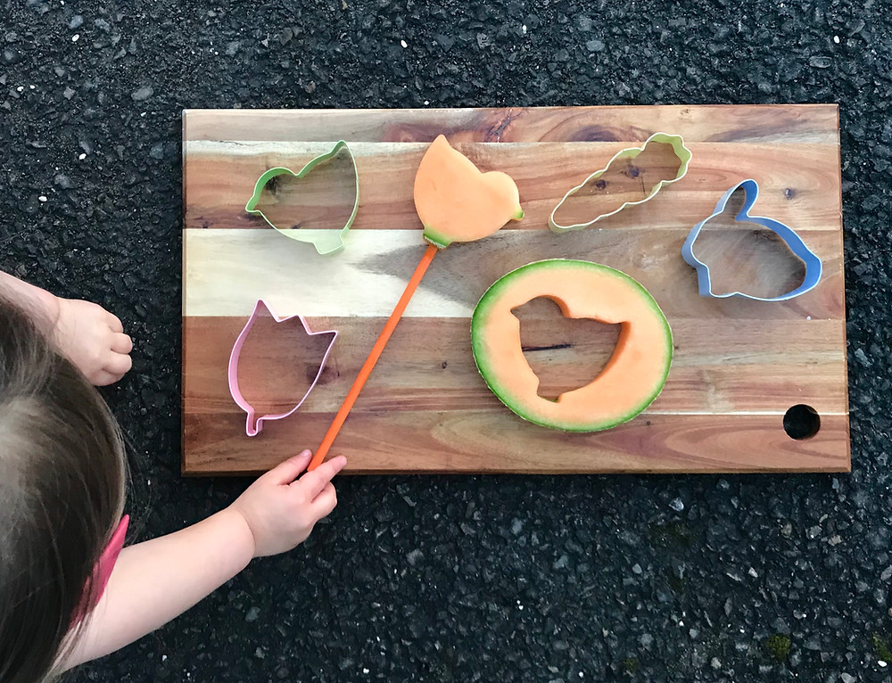 Cutting board show healthy cut outs of melon in Easter-themed cut-outs in the shape of bunnies, flowers and chicks. Cookie-cutters easily turn fruit into fun shapes