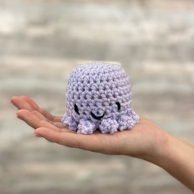Crocheted lilac colour octopus with a smiley face, sitting on an open hand.