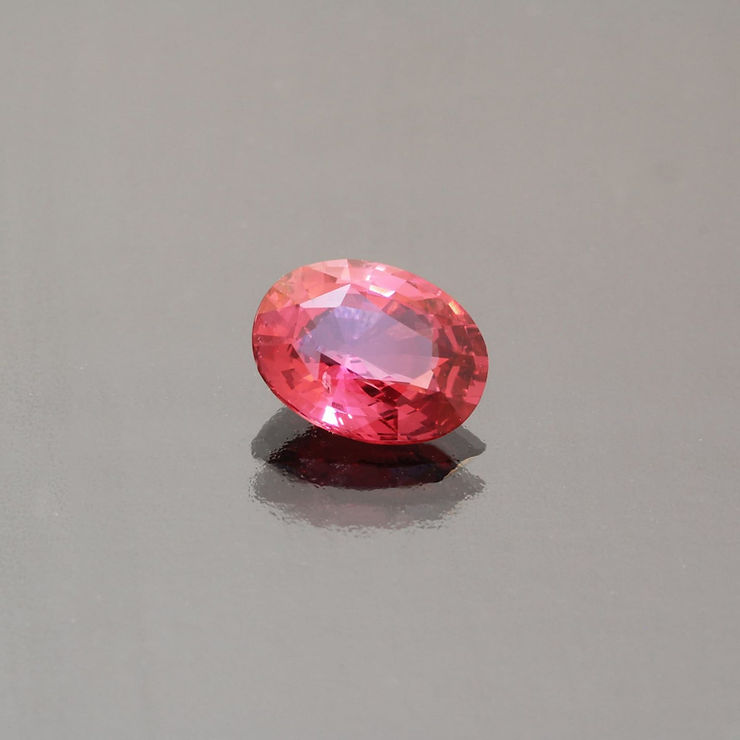 Ein rosa Spinell-Edelstein mit facettiertem Ovalschliff liegt auf einer reflektierenden grauen Oberfläche und erzeugt einen subtilen Schatten.
