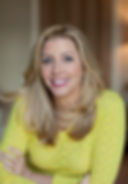Smiling woman with long blonde hair wears a bright yellow sweater. She has her arms crossed and is in a warmly lit, blurred background.