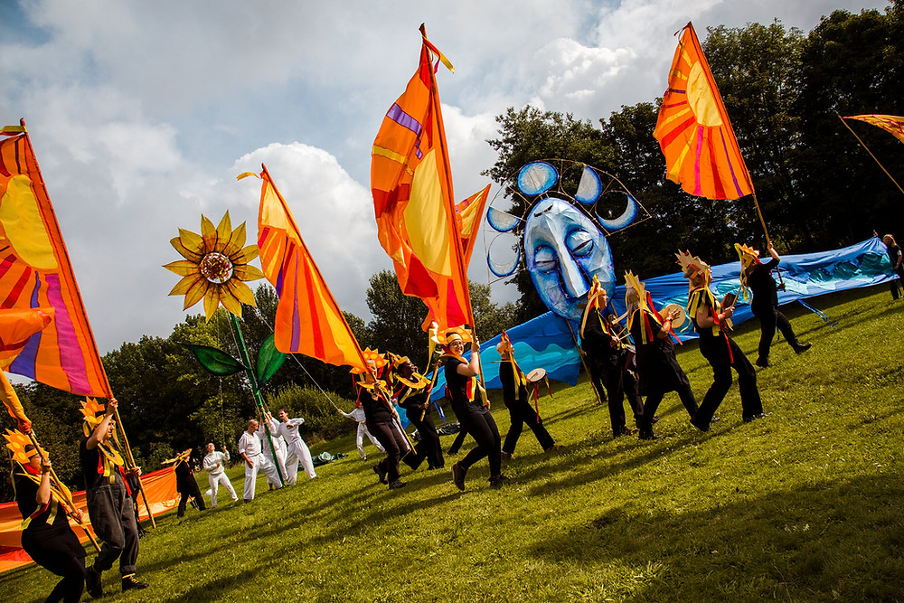 Lots of performers walk across a green field holding orange flags. In the background a giant yellow sunflower and giant blue moon puppet stand.