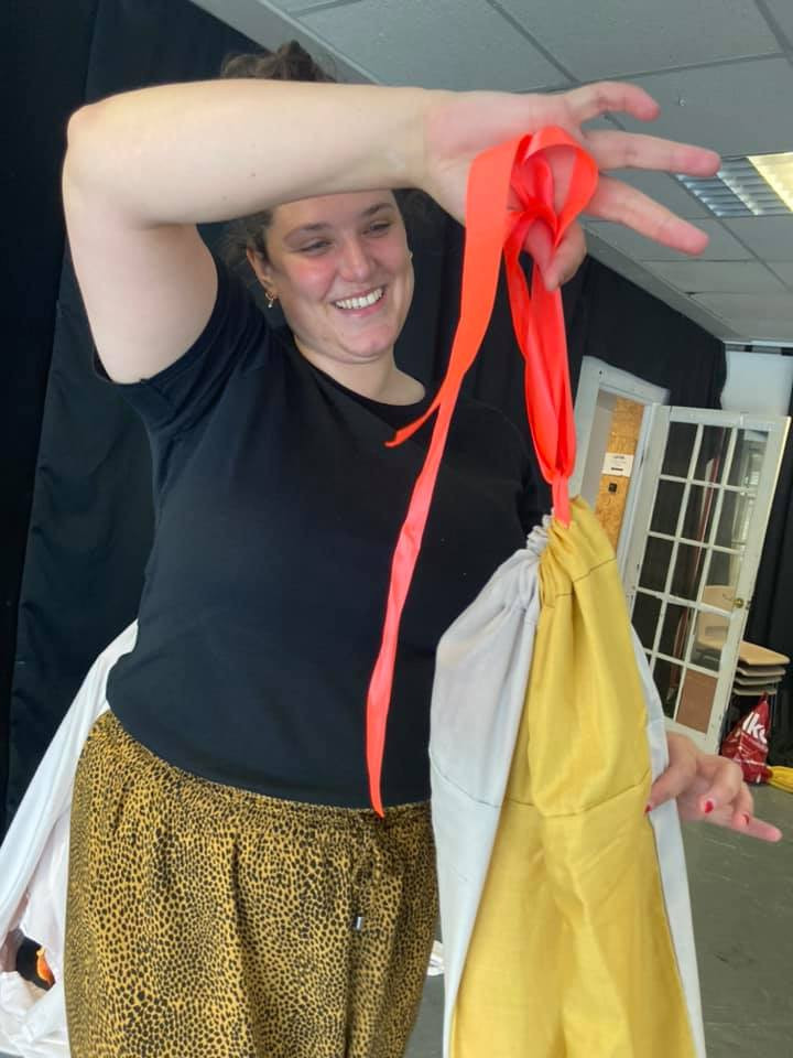 A smiling white woman with brown hair holds up a yellow and white material bag with an orange ribbon which she has just sewn