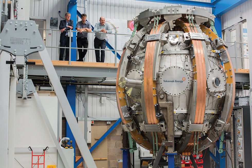 Three people observe a large fusion device labeled Tokamak Energy in an industrial setting. The device is metallic with copper details.