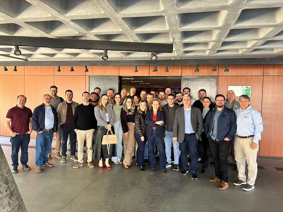 Foto em grupo com cerca de 30 profissionais, entre homens e mulheres, sorrindo e posicionados lado a lado em um ambiente corporativo ou de eventos, com piso de cimento queimado, teto com estrutura de concreto aparente em formato quadriculado e painéis de madeira ao fundo.