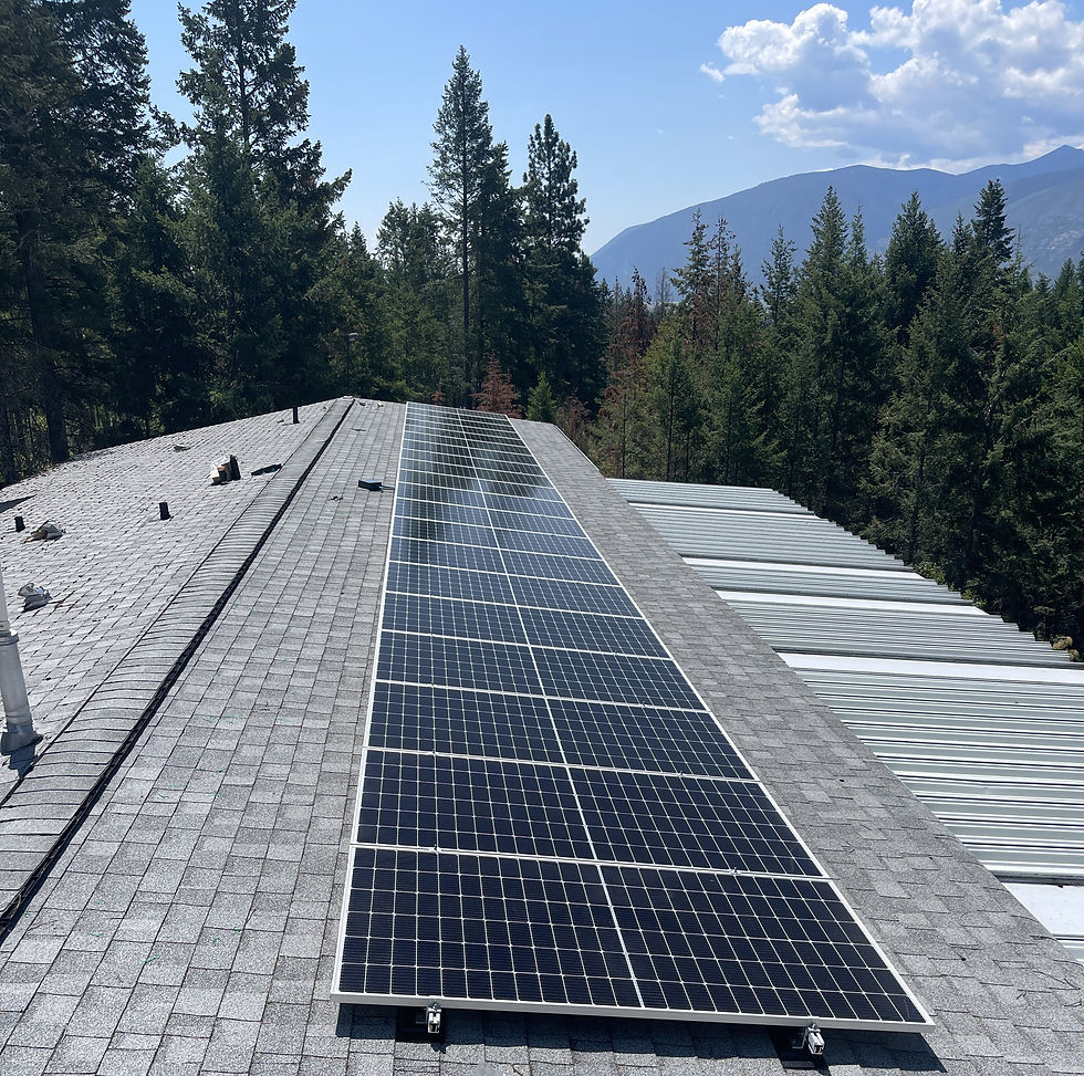 Solar panels on a gray shingled roof surrounded by pine trees and mountains under a partly cloudy sky.