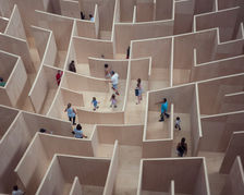 People navigating a large wooden maze, viewed from above. The maze has tall, light brown walls, and participants appear focused and curious.