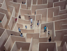 People navigating a large wooden maze, viewed from above. The maze has tall, light brown walls, and participants appear focused and curious.
