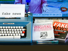 Blue typewriter with "fake news" paper, books on digital verification and fake news beside it. TV in the background shows journalist.