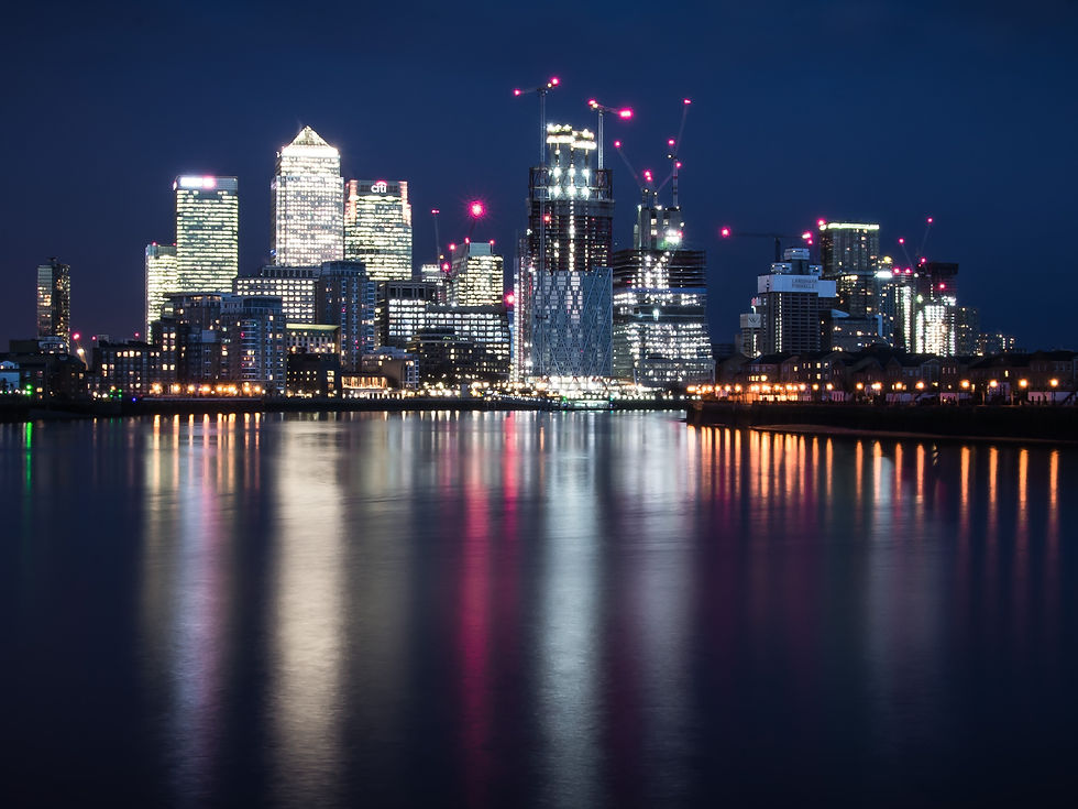 City skyline at night with tall buildings, blurred red and white lights from traffic on a highway in the foreground, against a cloudy sky.