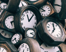 Cluster of vintage wall clocks with white faces and black hands, piled together in a rustic, moody close-up.