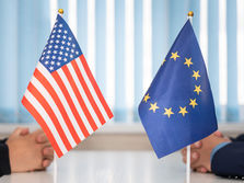 U.S. and EU flags on a table, hands clasped in the background. Blue vertical blinds set a formal tone, highlighting international dialogue.
