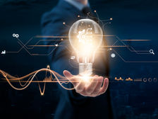 A person holds a glowing light bulb with digital symbols and graphs floating around, suggesting innovation. Background is dark and urban.