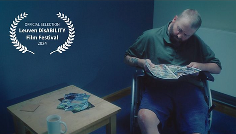 A white man with short hair sits in a wheelchair holding a magazine. There is a cuppa and some paper