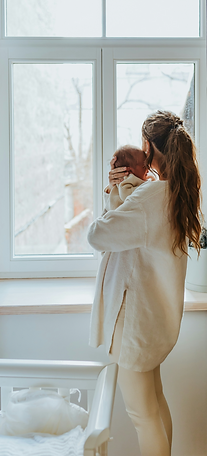 Mother cradles newborn, standing by a window, near a white crib, in daylight.