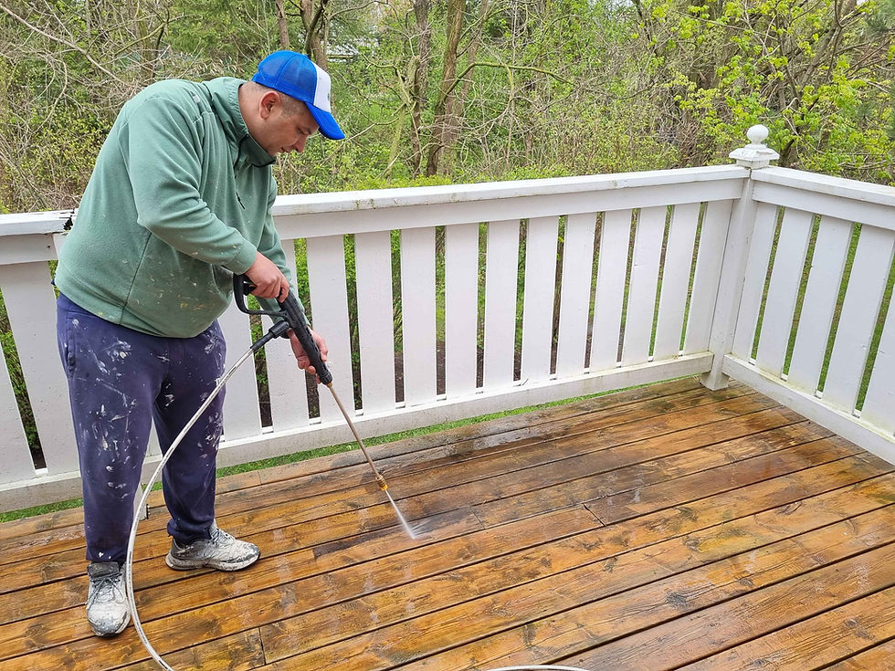 painter power washing the surface of a wooden deck with white railings and trees in the background