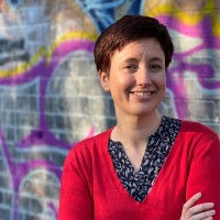 Smiling woman in a red sweater stands with arms crossed before a colorful graffiti wall.