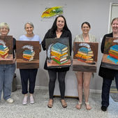 a group of women holding up their acrylic paintings of a stack of books with a feather quill on top.