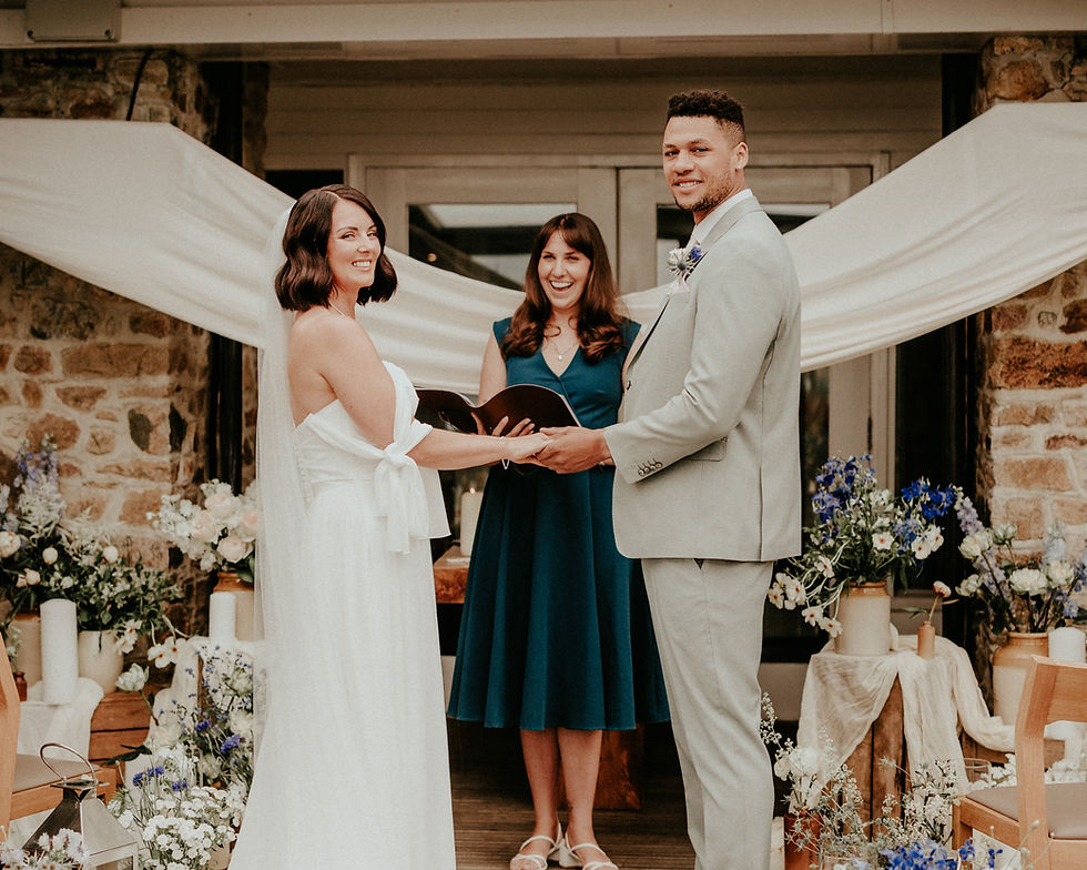 Bride and groom holding hands, smiling at each other, with officiant behind them. Stone background, floral decor, and white drapery.