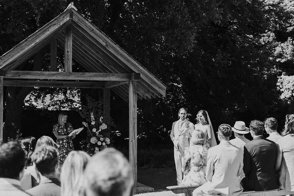Black-and-white outdoor wedding ceremony under a wooden pavilion, with the bride and groom facing guests and an officiant reading