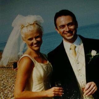 A photo of a couple just married in the 1990s - they are on the beach each holding a glass of fizz - she is wearing a gold coloured dress with a  short veil and he is in a tailsuit with a gold waistcoat
