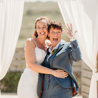 Two brides are standing under voiles at an outdoor ceremony - one in a white dress and the other a pale blue suit. They are grinning and one in holding her hand aloft, showing off her wedding ring