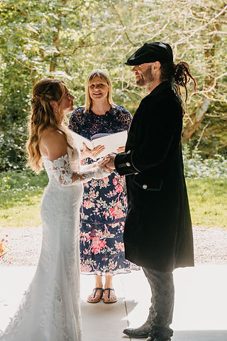 A wedding celebrant in a summery dress officiates a wedding at a rustic barn - the couple stand either side of her
