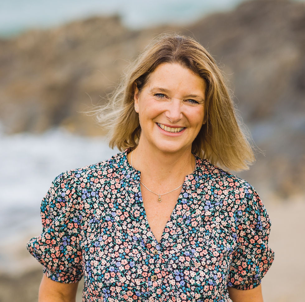 Smiling woman in a floral dress stands on a rocky beach. Her hair is windswept, creating a cheerful, relaxed atmosphere.