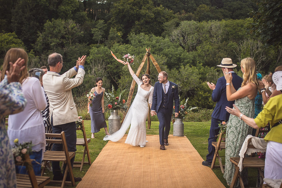 Bride and groom joyfully walk down aisle, bride waving bouquet. Guests clap in outdoor setting with lush greenery. Festive and celebratory.