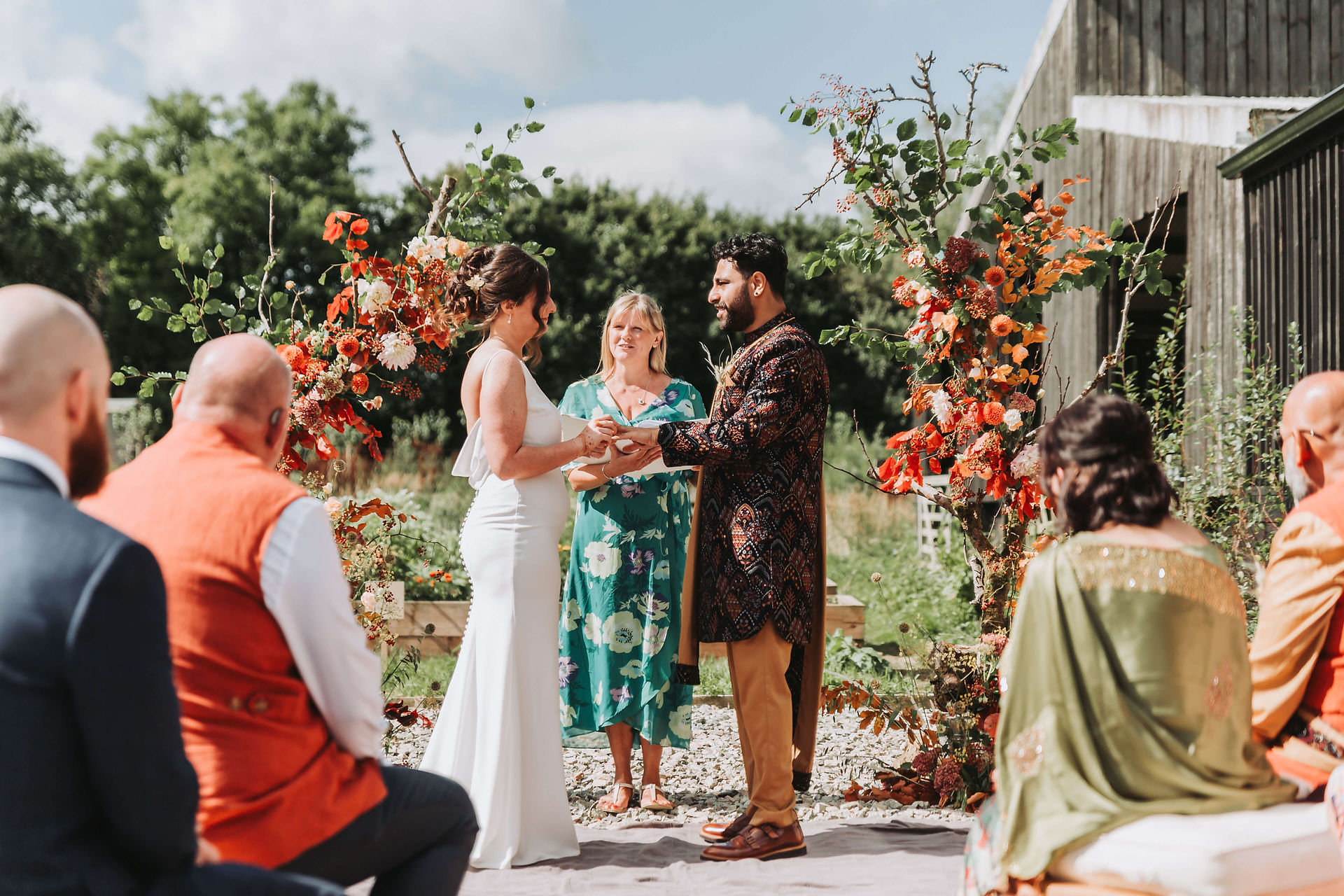 Celebrant in a green patterned dress stands between bride and groom at British-Indian fusion wedding ceremony