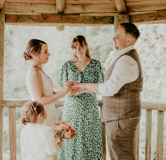 A celebrant looks to camera in a wooden pergola with a bride and groom and small girl holding a posy