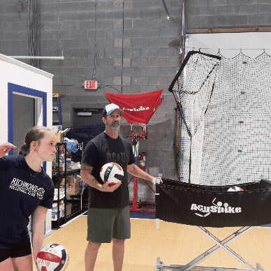 A girls volleyball player practicing her serving with a coach nearby.