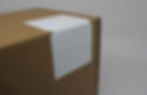 Brown cardboard box close-up with a blank white label on its corner against a plain background. Simple and neutral setting.