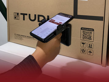 Hand holds a phone scanning a large brown TUDI box. Visible text includes TUDI logos and website, with a QR code and symbols on the box.