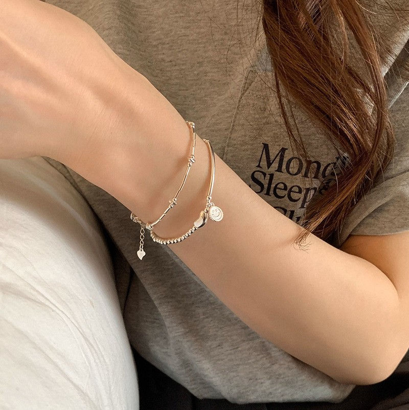 A close-up image of a silver beaded bracelet with a smiley face charm and a curved silver bar, held by a hand with manicured