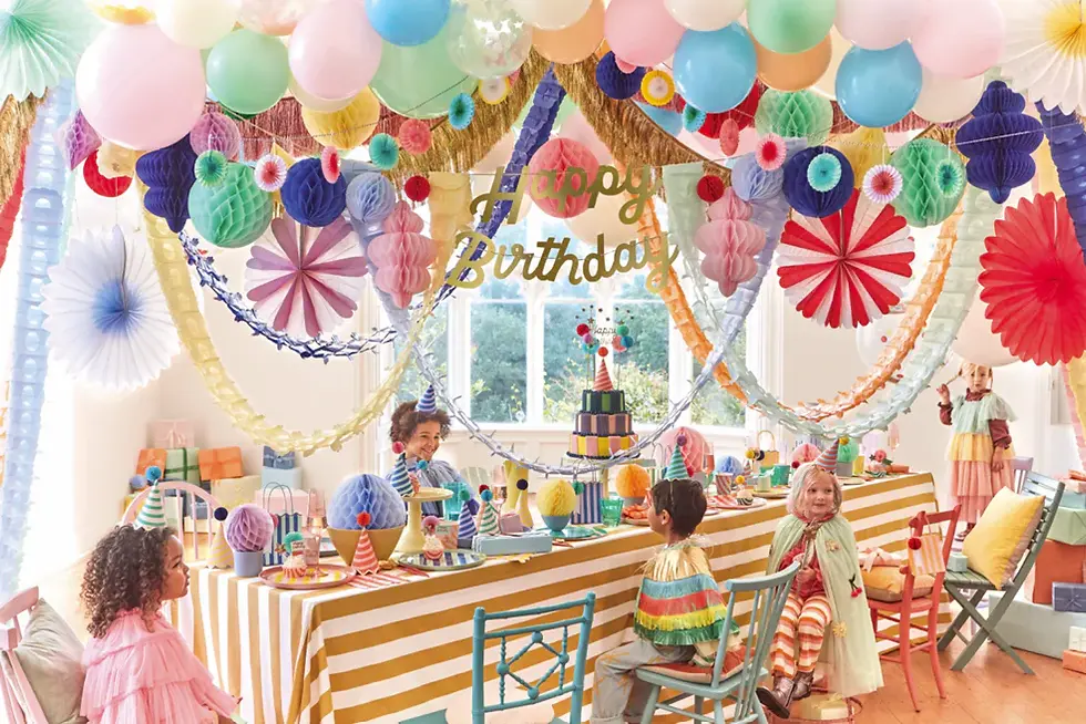 Kids at a colorful birthday party with balloons, striped tablecloth, cake, and "Happy Birthday" banner in a bright room full of decorations.