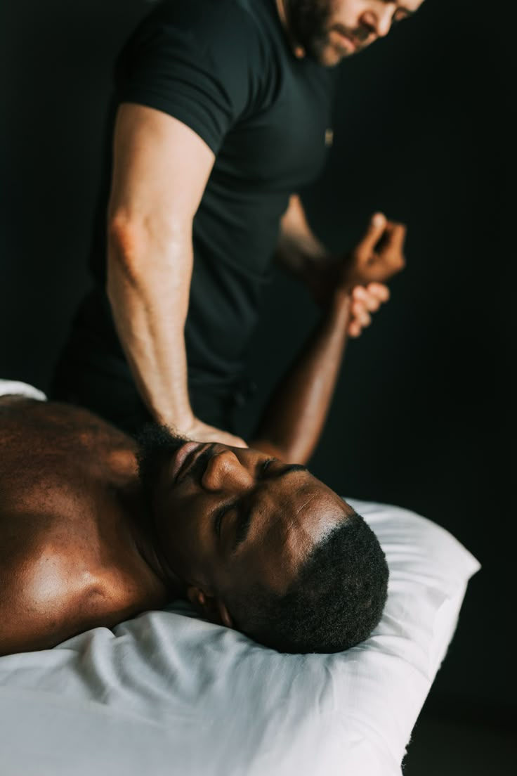 Man in black shirt giving a shoulder massage to a relaxed man lying on a white pillow. Dimly lit, serene atmosphere.