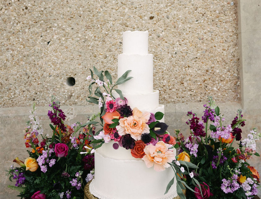 A five-tier white wedding cake with textured frosting and cascading fresh flowers including orange roses and purple snapdragons, set against a stone wall background.