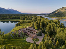 Aerial view of a large house surrounded by lush trees, a river, and a lake. Mountains in the background under a clear blue sky. Tranquil setting.