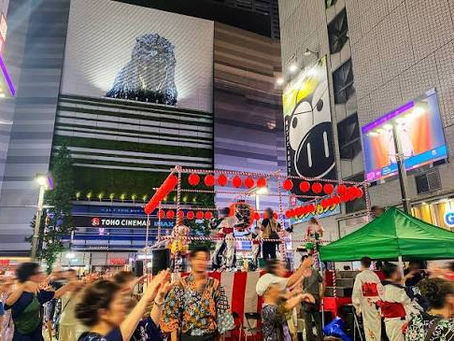 東京新宿歌舞伎町夜間夏祭活動中，Cinecity 廣場設有盆舞高台，周邊被霓虹燈、高樓與人群包圍，現場亦有哥斯拉立體像和表演舞台。