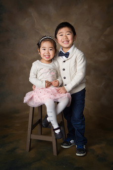 A smiling boy and girl are dressed in formal attire, sitting on a stool against a brown background.