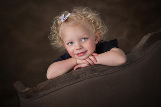 A smiling child with curly hair leans on a brown armchair, wearing a dark top and floral hairpin, se